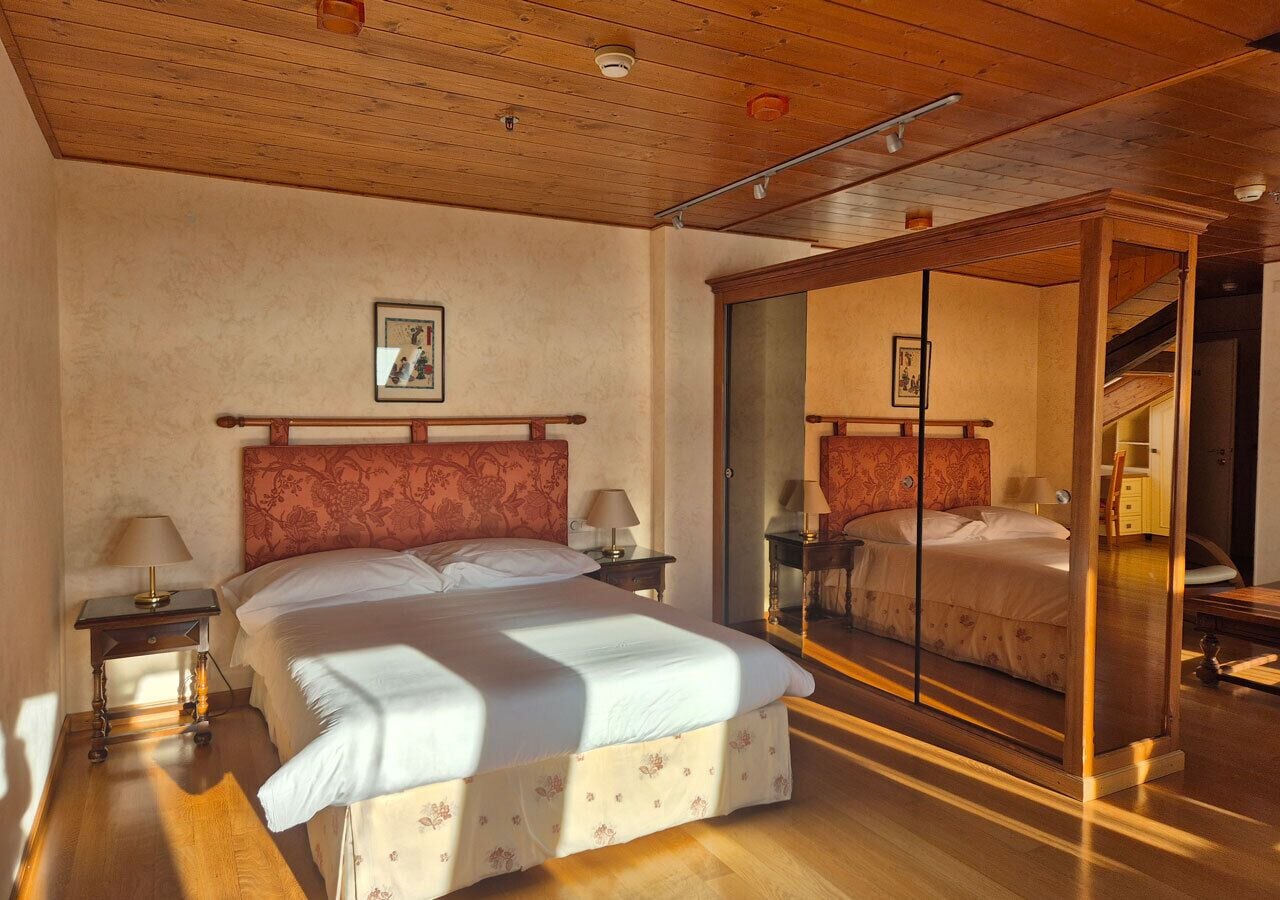 A bedroom with a large bed, a wooden ceiling, a mirror, and a nightstand with a lamp.