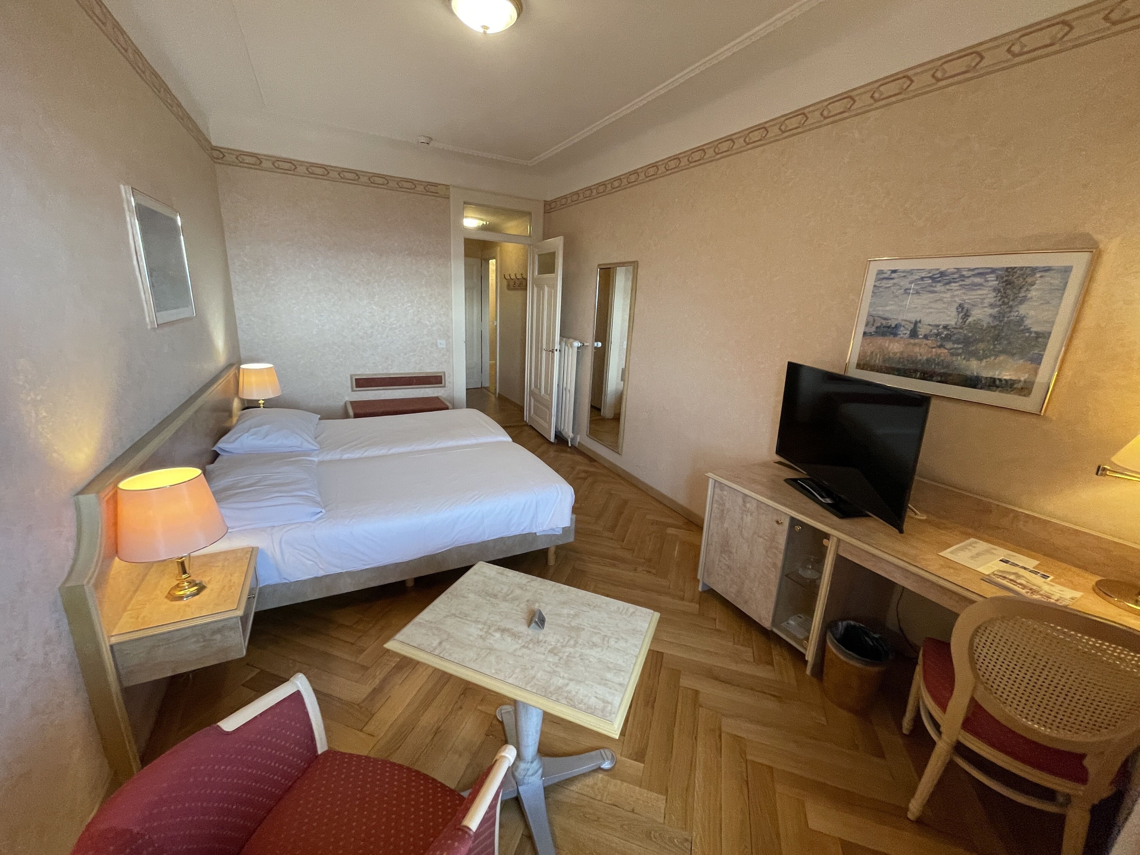 Una habitación de hotel con cama, escritorio con televisor, silla, mesita y espejo.