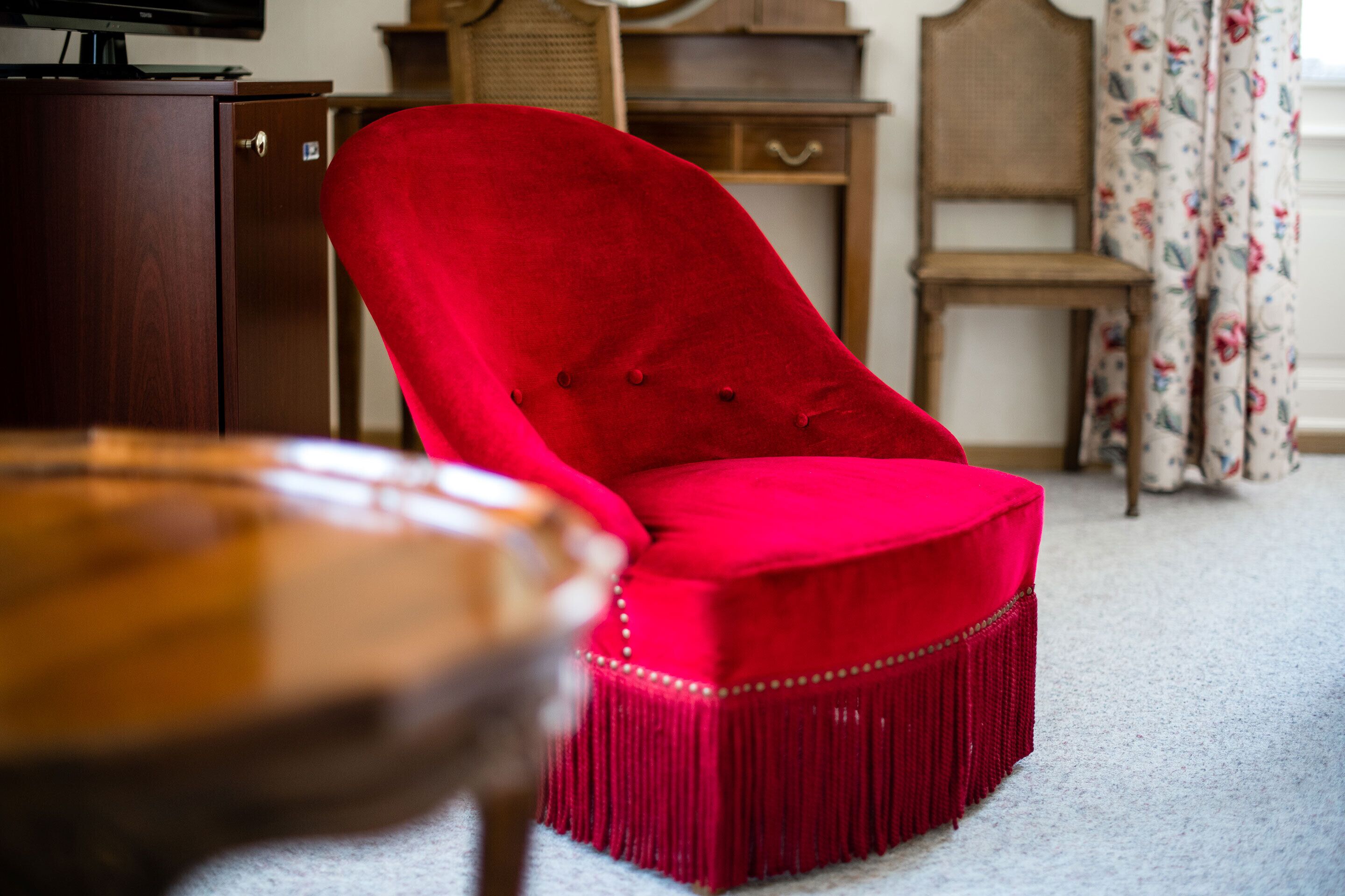 A red upholstered chair with a fringe trim, a wooden cabinet, and a floral curtain in a room with a carpeted floor.