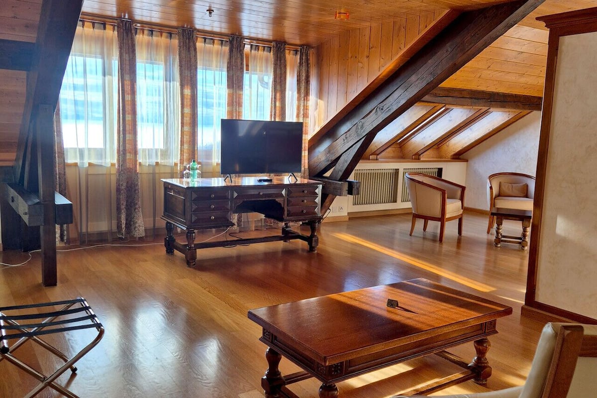 A spacious attic room with wooden floors, a large wooden desk, a television, and a dining table.