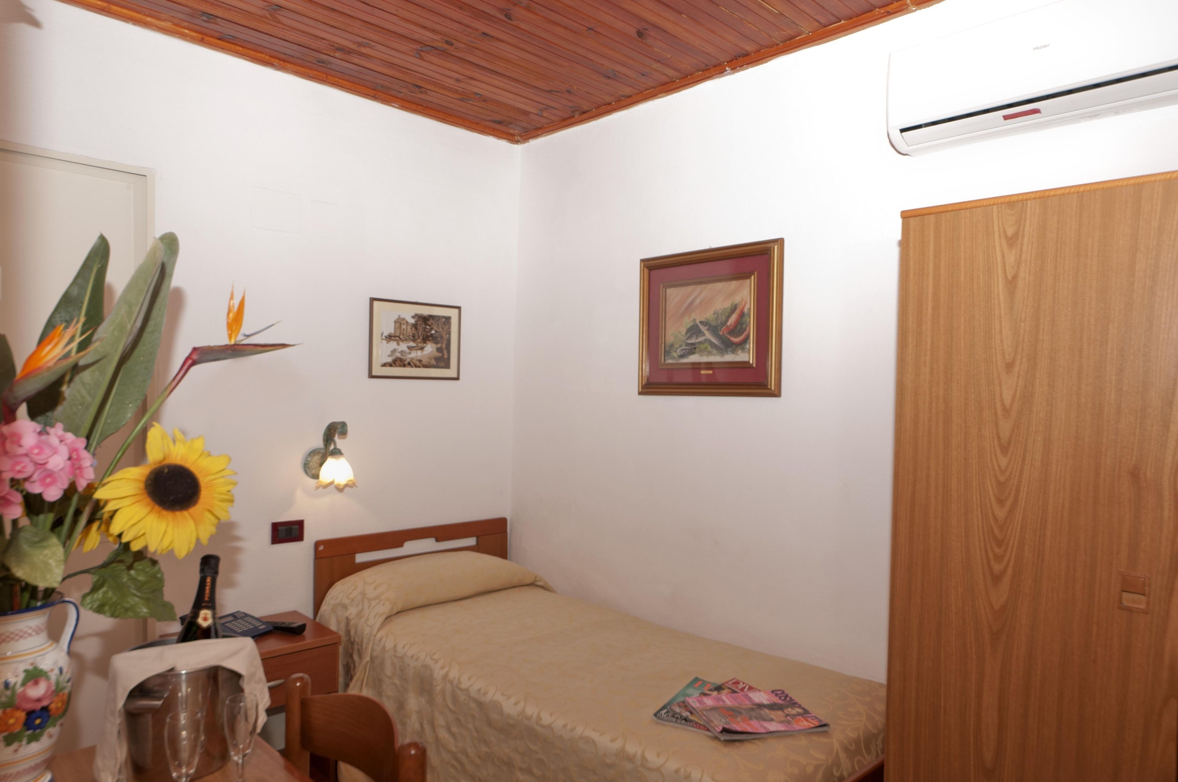 Habitació petita i ordenada amb un llit individual, un armari de fusta, aire condicionat fixat a la paret i una taula petita amb un ram de flors i una ampolla.