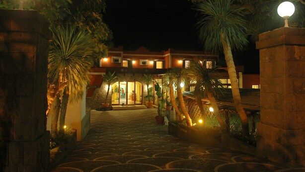 Entrada de un hotel de noche con un camino empedrado, palmeras y una fachada iluminada.