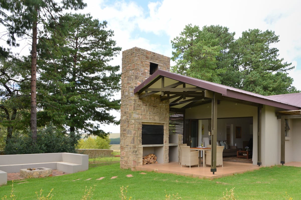 A modern outdoor patio area with a stone fireplace, seating, and a covered section with a door leading to a private room.