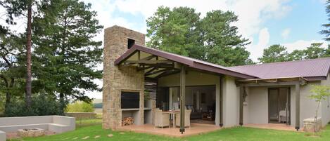 A modern outdoor patio area with a stone fireplace, seating, and a covered section with a door leading to a private room.