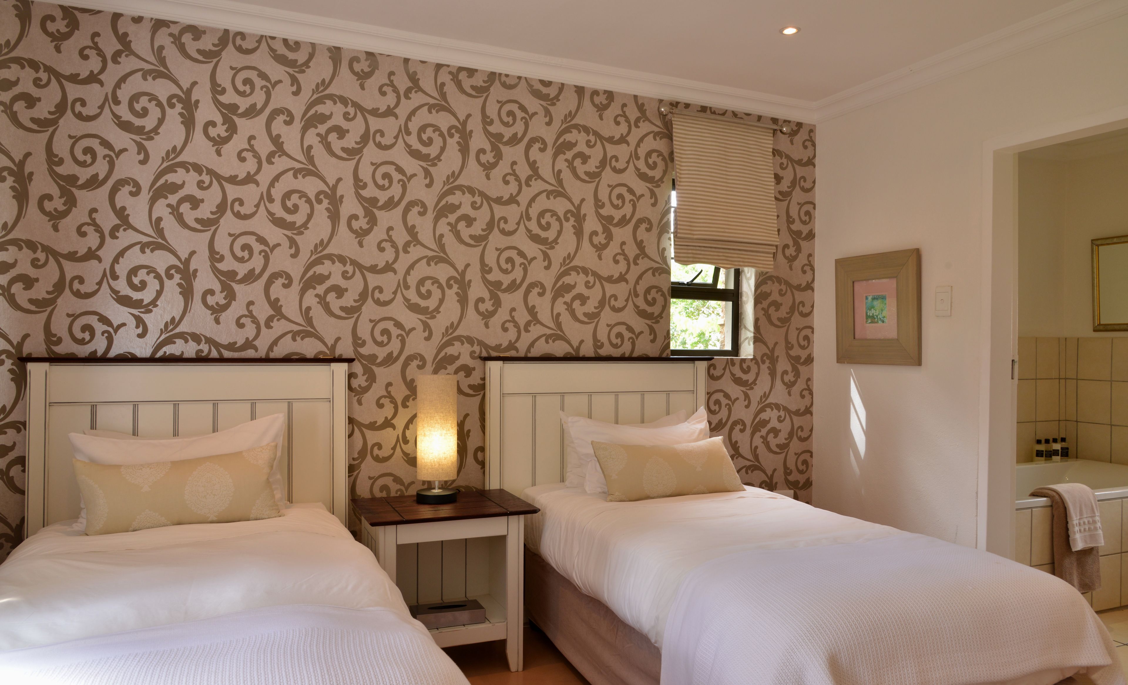 A bedroom with two beds, a nightstand with a lamp, patterned wallpaper, a window with blinds, and a framed picture on the wall.