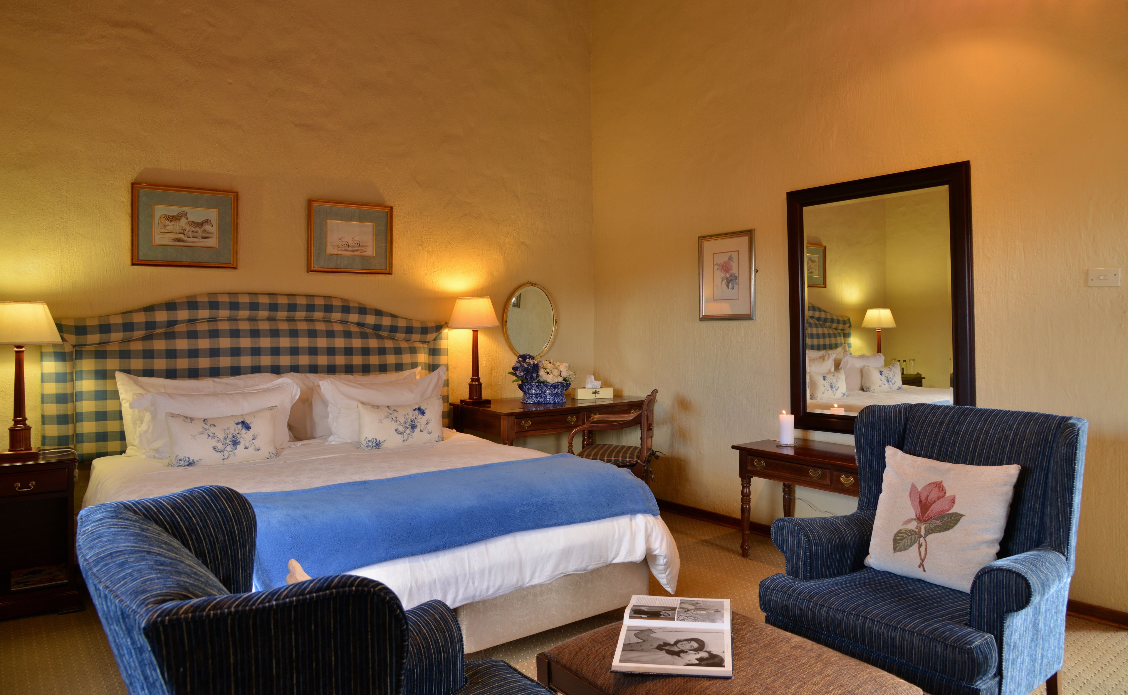 Habitación de hotel con cabecera a cuadros, cama grande con ropa de cama blanca y azul, zona de estar con dos sillones, mesita de madera con lámpara y un espejo en la pared.