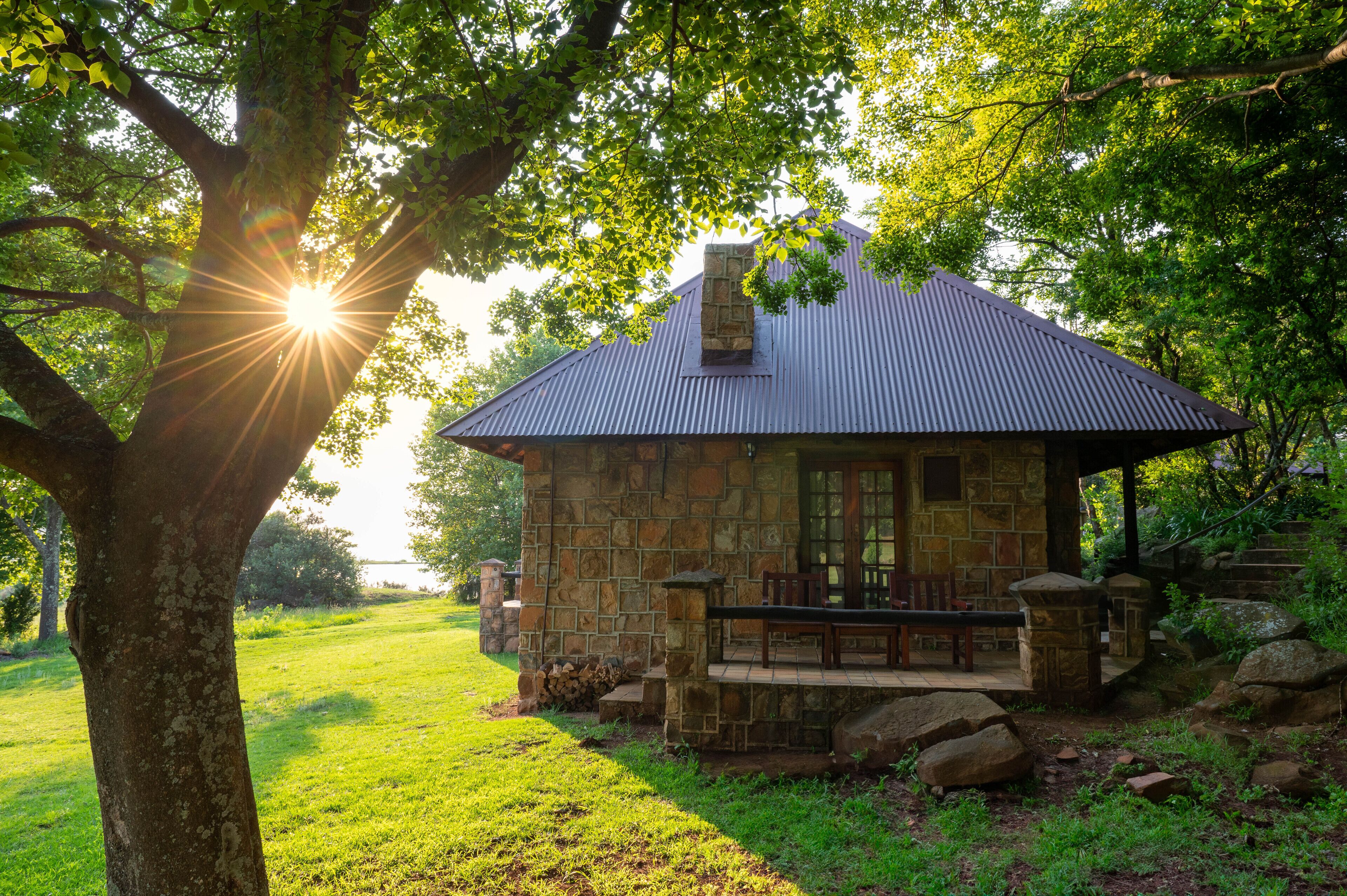 A stone cabin with a metal roof, surrounded by trees and a grassy area.