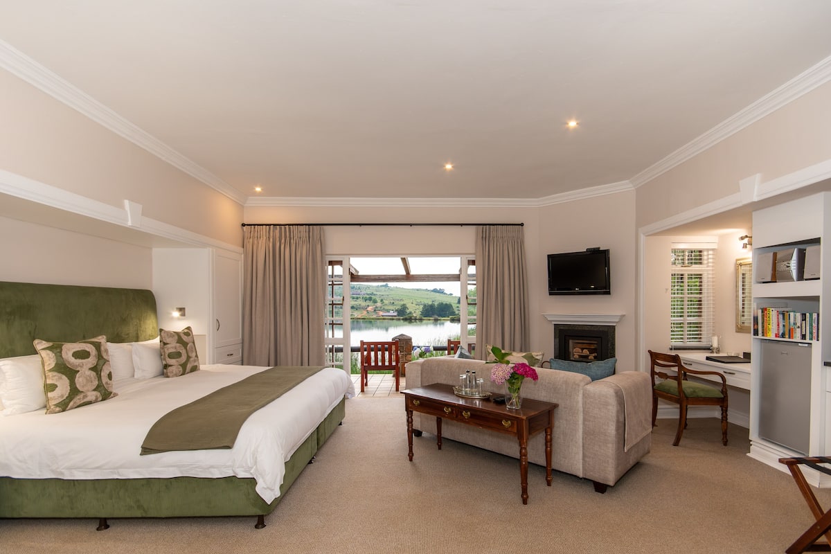 A bedroom with a large bed, a sofa, a desk, and a TV. There is a window with a view of a lake.
