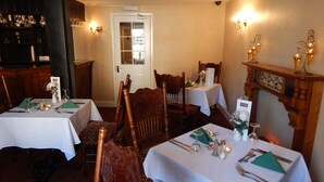 Un comedor con mesas dispuestas para cuatro, cada una con mantel blanco, cubiertos y servilletas verdes. Hay sillas de madera y un mueble de madera con copas.