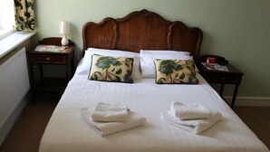 Una cama bien tendida con sábanas blancas y dos almohadones decorativos, una mesita de noche con lámpara, y una ventana con cortinas.