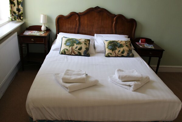 Una cama bien tendida con sábanas blancas y dos almohadones decorativos, una mesita de noche con lámpara, y una ventana con cortinas.