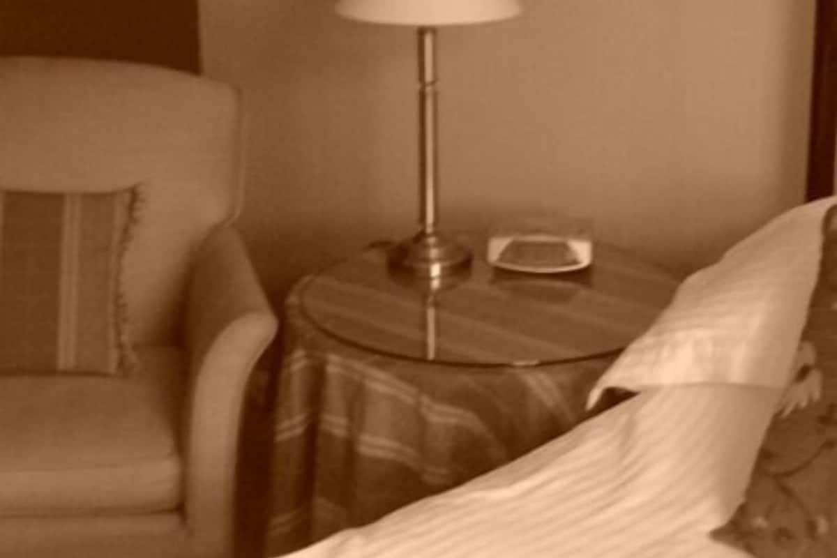 Quarto de hotel com uma cama, uma cadeira, um abajur e uma pequena mesa com um rádio.