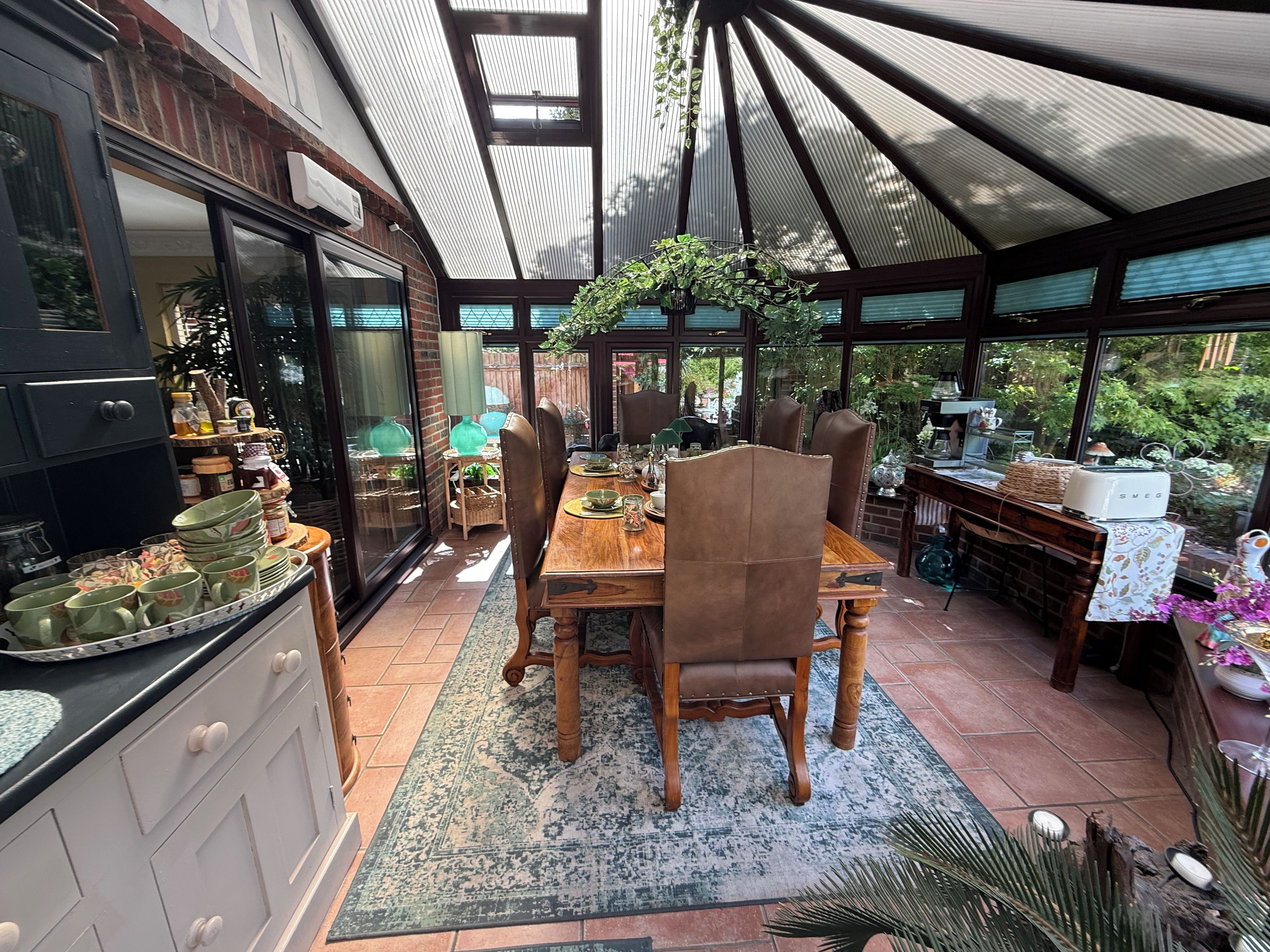 Ein Essbereich mit Holztisch und -stühlen, eine Küche mit Schränken und einem Kühlschrank sowie eine Terrasse mit einer Pergola und Pflanzen.