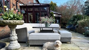 Un patio con un sofá de mimbre, una maceta de piedra y un perro pequeño.
