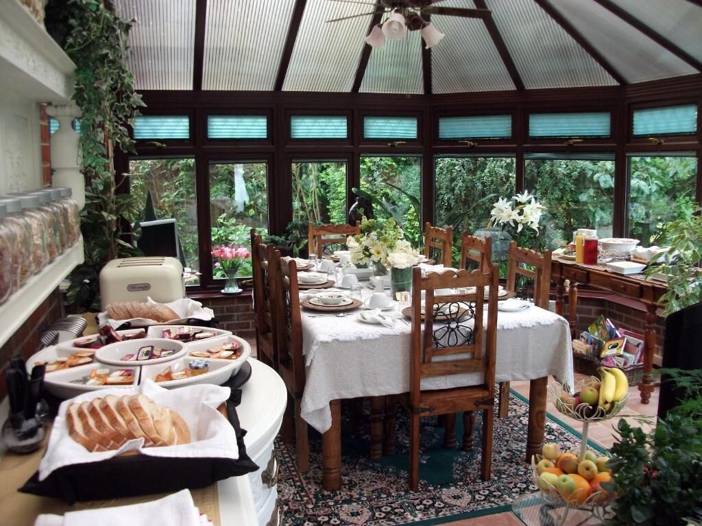 A conservatory dining area with a table set for a meal, surrounded by chairs, and a fruit basket on the side.