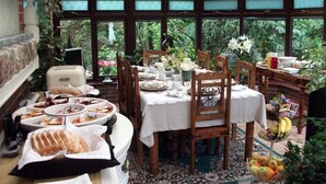 A conservatory dining area with a table set for a meal, surrounded by chairs, and a fruit basket on the side.