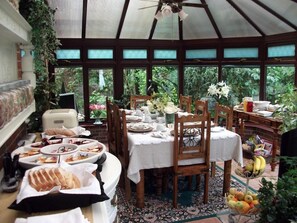 A conservatory dining area with a table set for a meal, surrounded by chairs, and a fruit basket on the side.
