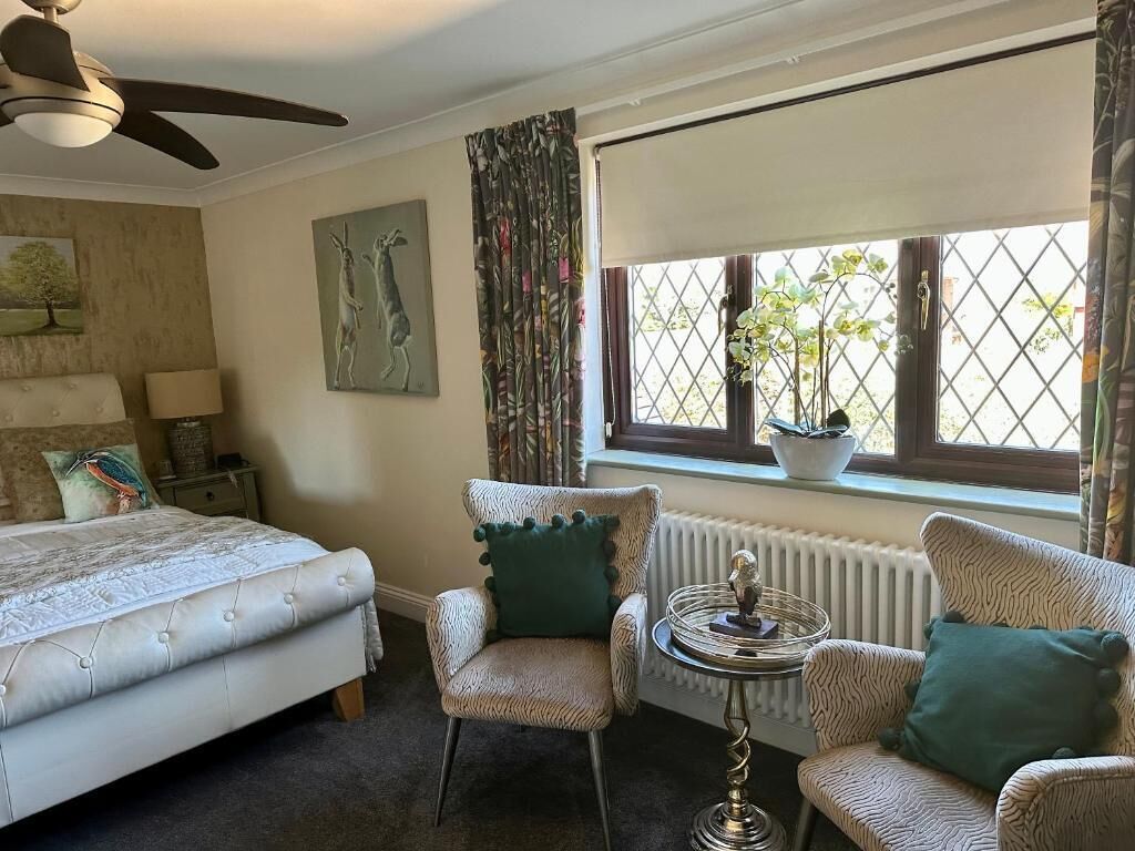 Un dormitorio con una cama, dos sillones, una mesita, un ventilador de techo, una ventana con cortinas y un radiador.