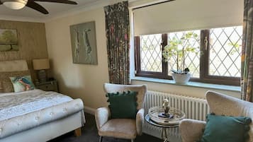 Un dormitorio con una cama, dos sillones, una mesita, un ventilador de techo, una ventana con cortinas y un radiador.