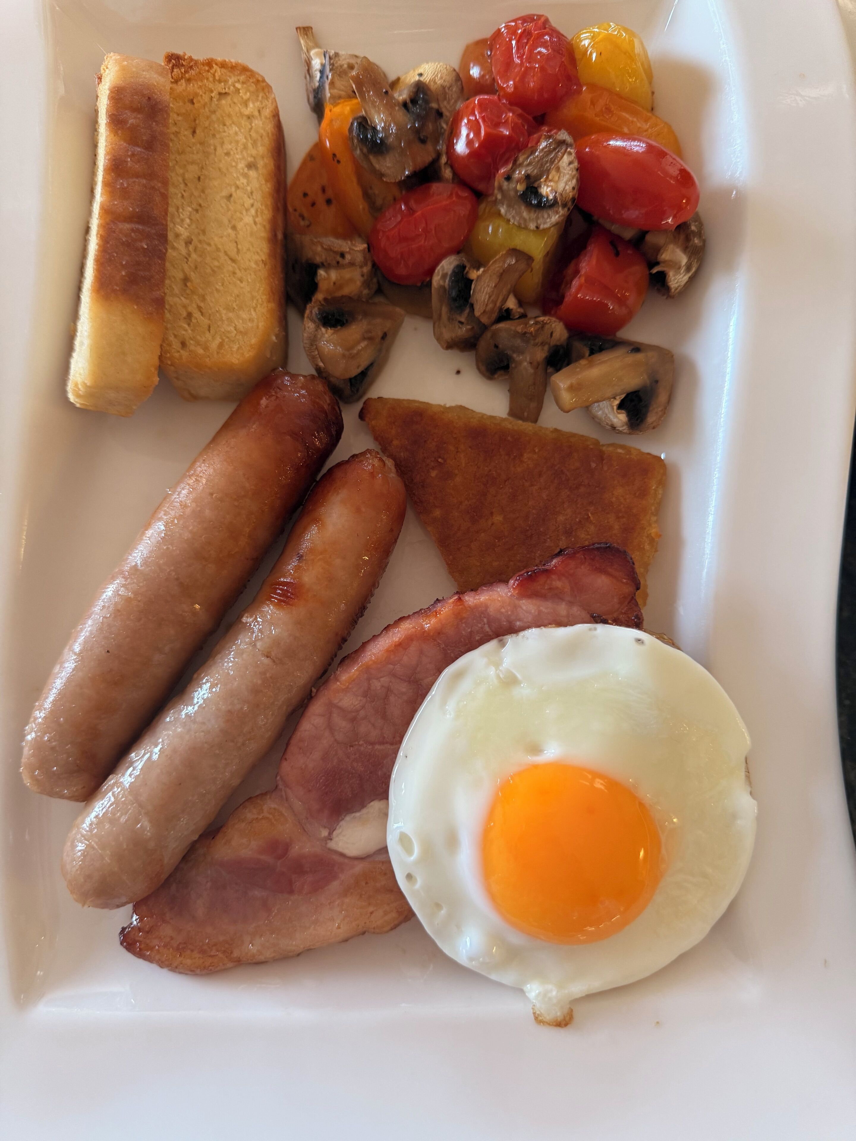 Une assiette de petit-déjeuner comprenant des toasts, des saucisses, du bacon, un œuf au plat, ainsi que des tomates et des champignons rôtis.