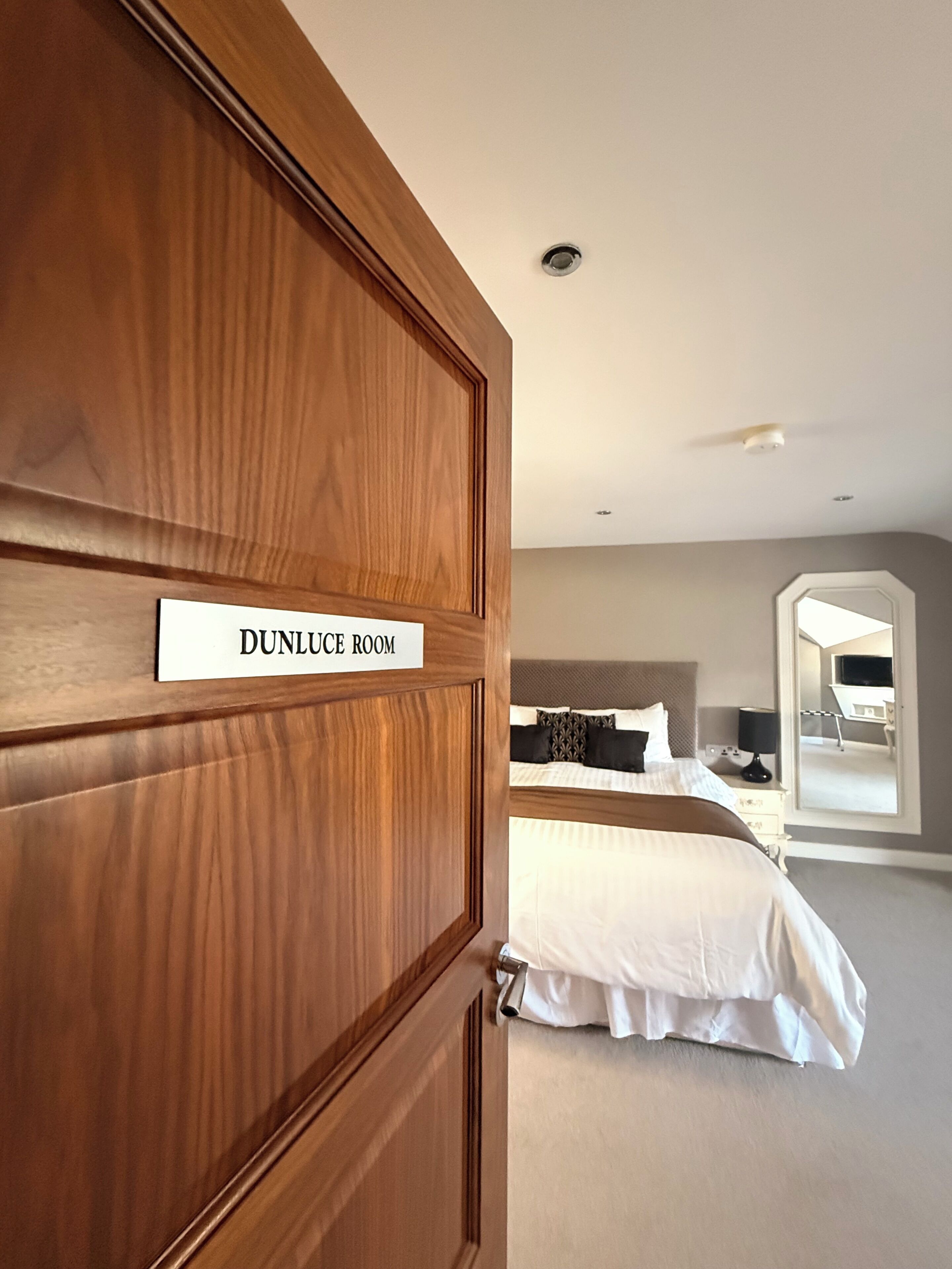 A hotel room with a wooden door labeled 'DUNLUC ROOM,' a bed with white bedding, a bedside table with a lamp, and a large mirror on the wall.