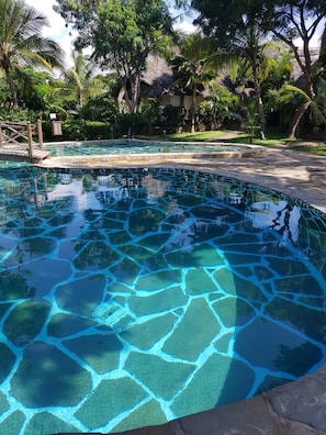 A clear pool with a stone pattern, surrounded by lush greenery and a thatched-roof structure.