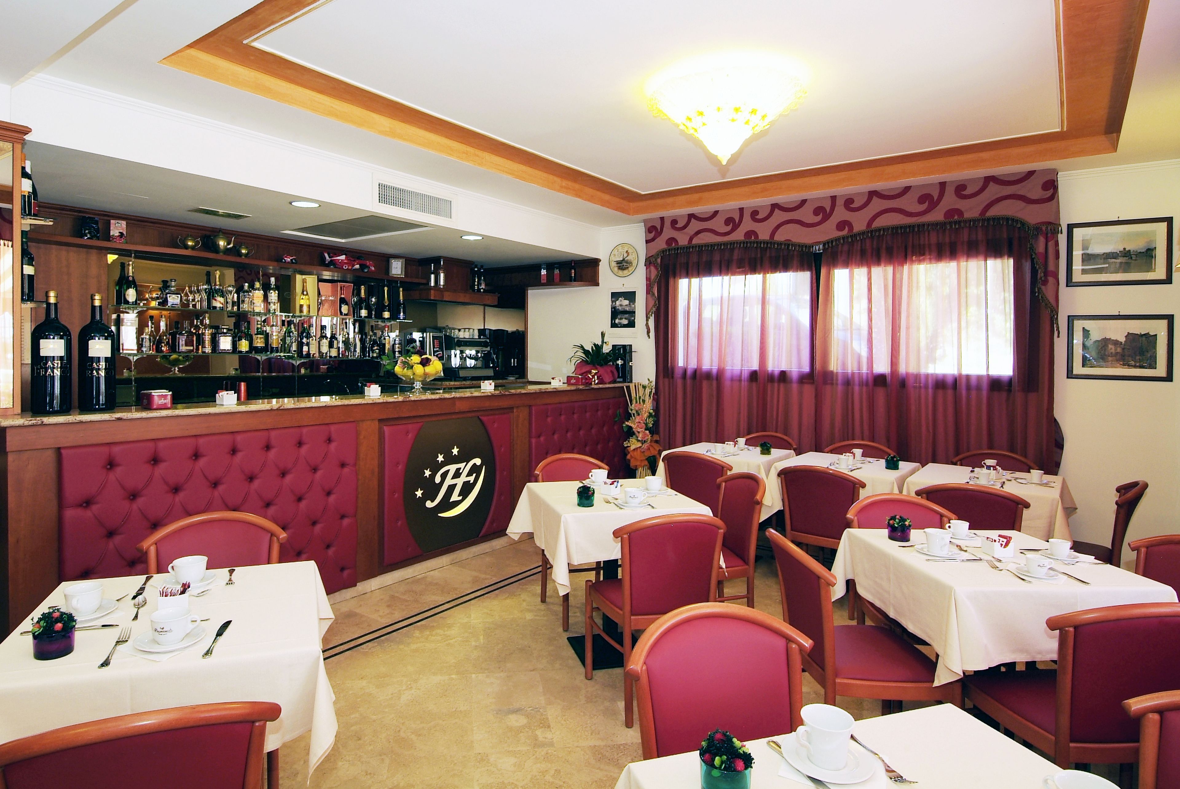A restaurant with multiple tables and chairs, a bar area with various bottles, and a decorative wall with a logo.
