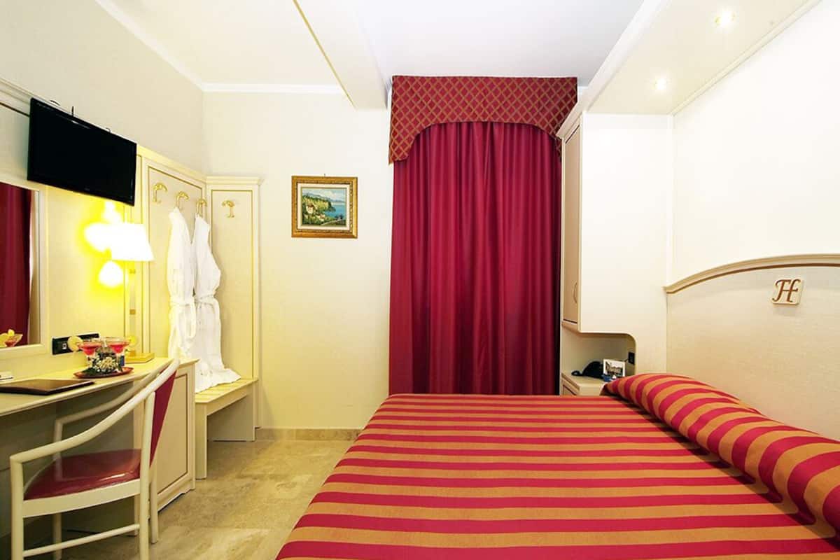 A hotel room with a bed, a desk with a chair, a television, and a red curtain.