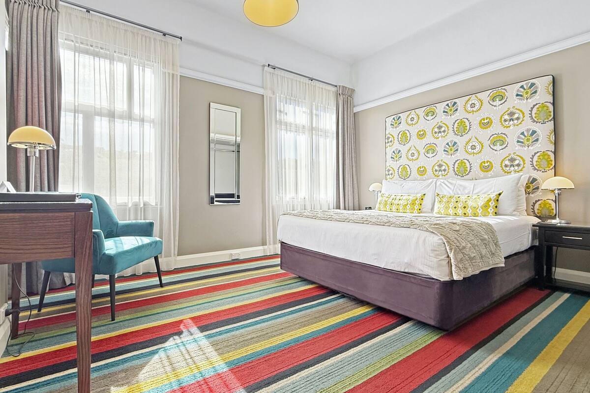 A hotel room with a bed, a desk, a chair, a colorful striped carpet, and a large decorative headboard.