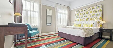 Une chambre d’hôtel comprenant un lit, un bureau, une chaise, une moquette rayée aux couleurs vives et une grande tête de lit décorative.