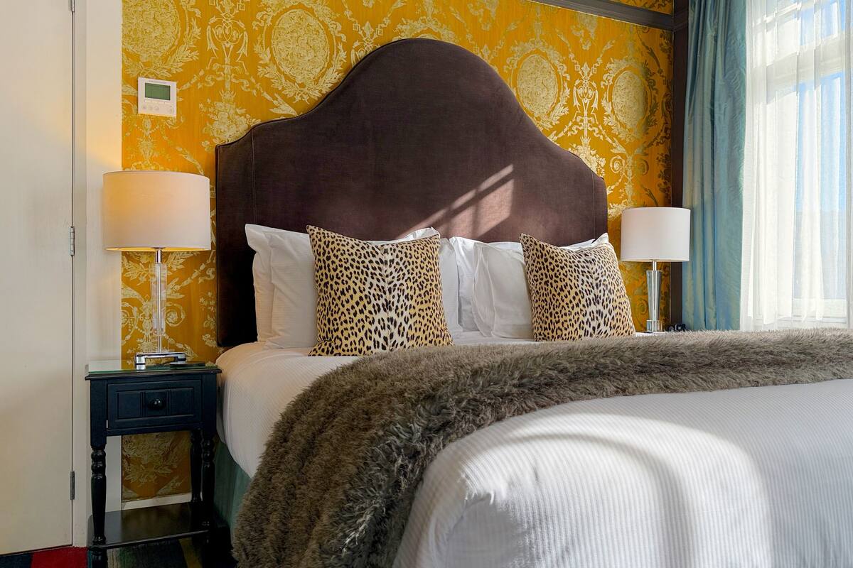 A hotel room with a large bed, two leopard print pillows, a fluffy throw, a nightstand with a lamp, and a thermostat on the wall.
