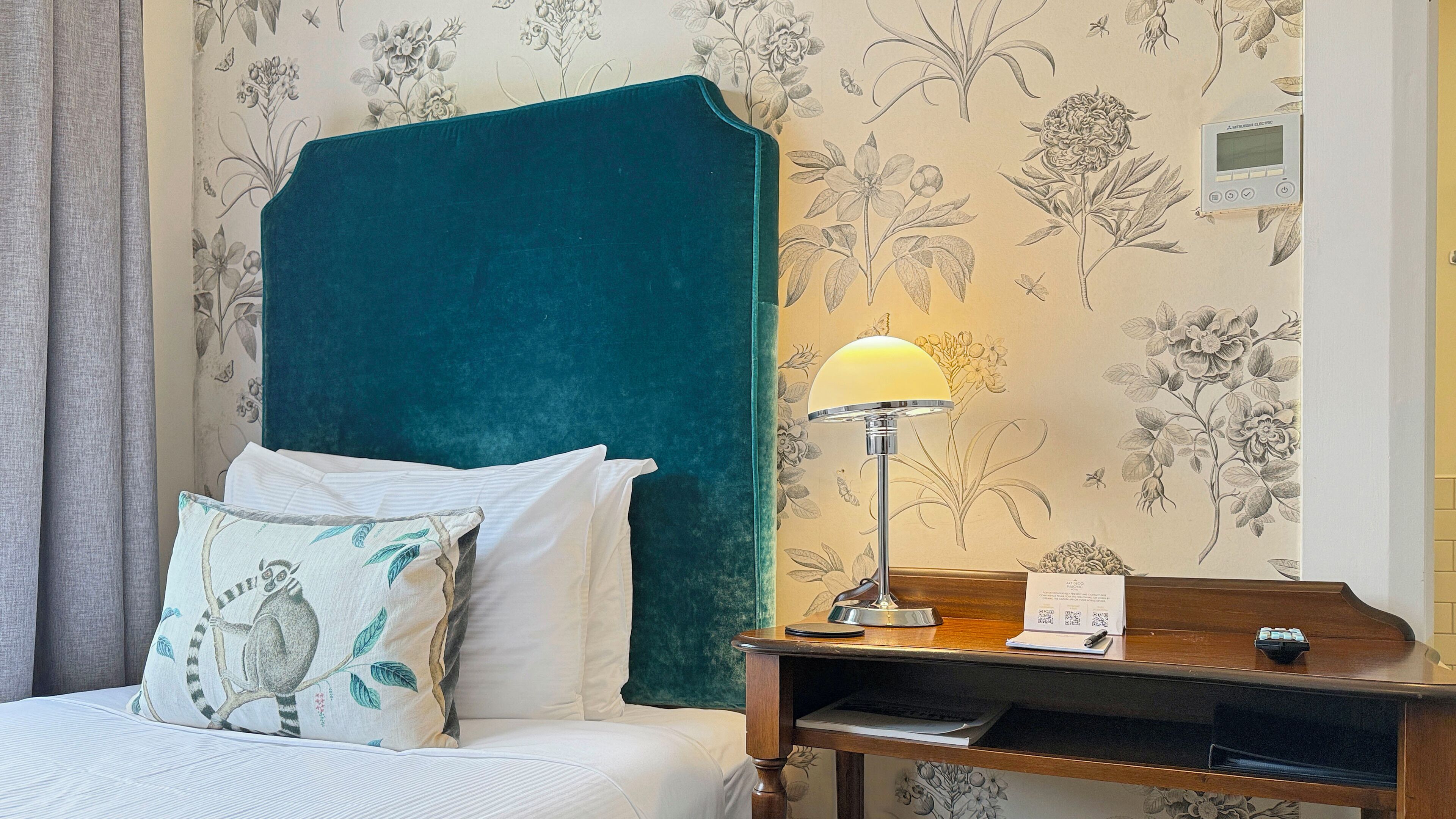 Una cama bien hecha con un cabecero color turquesa, una lámpara de noche y un papel tapiz floral.