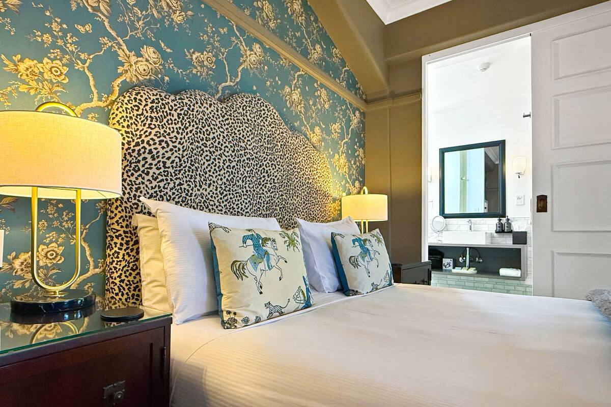 A hotel room with a bed, leopard print headboard, floral wallpaper, two bedside lamps, and a view of the bathroom.