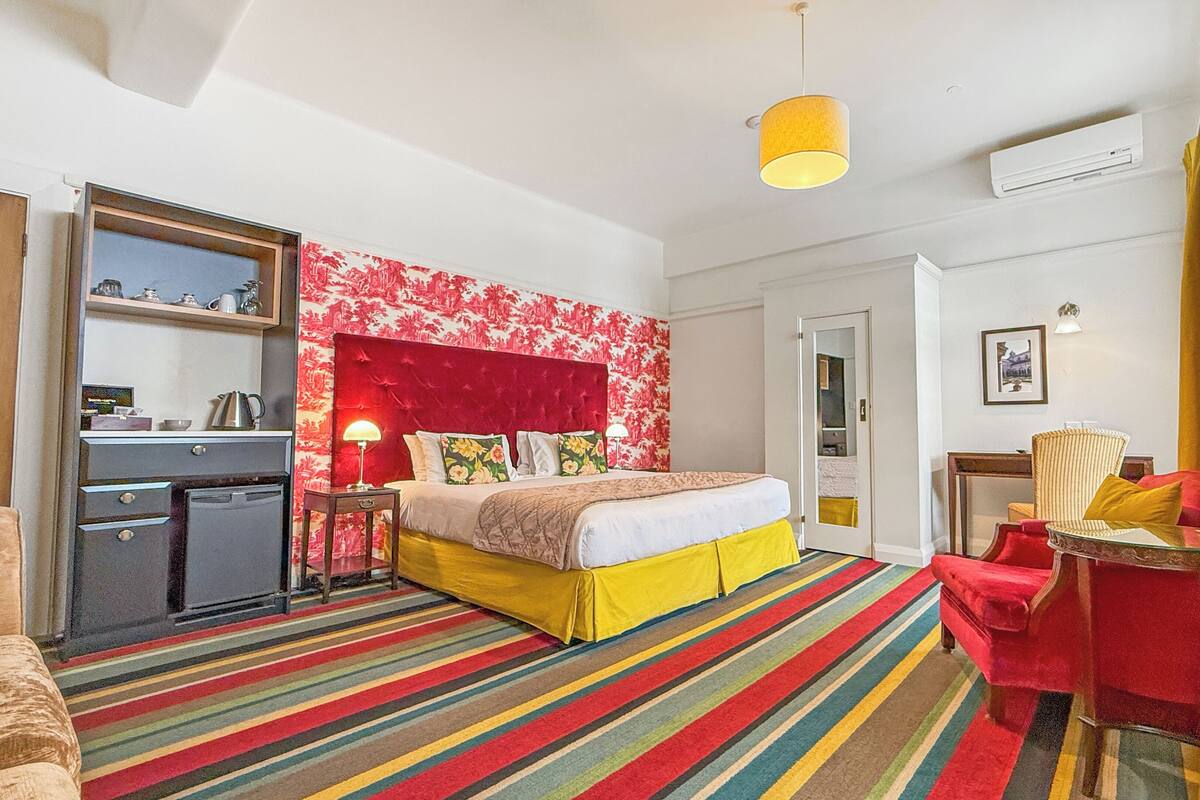 A hotel room with a bed, a desk, a chair, a small kitchenette, and a colorful striped carpet.