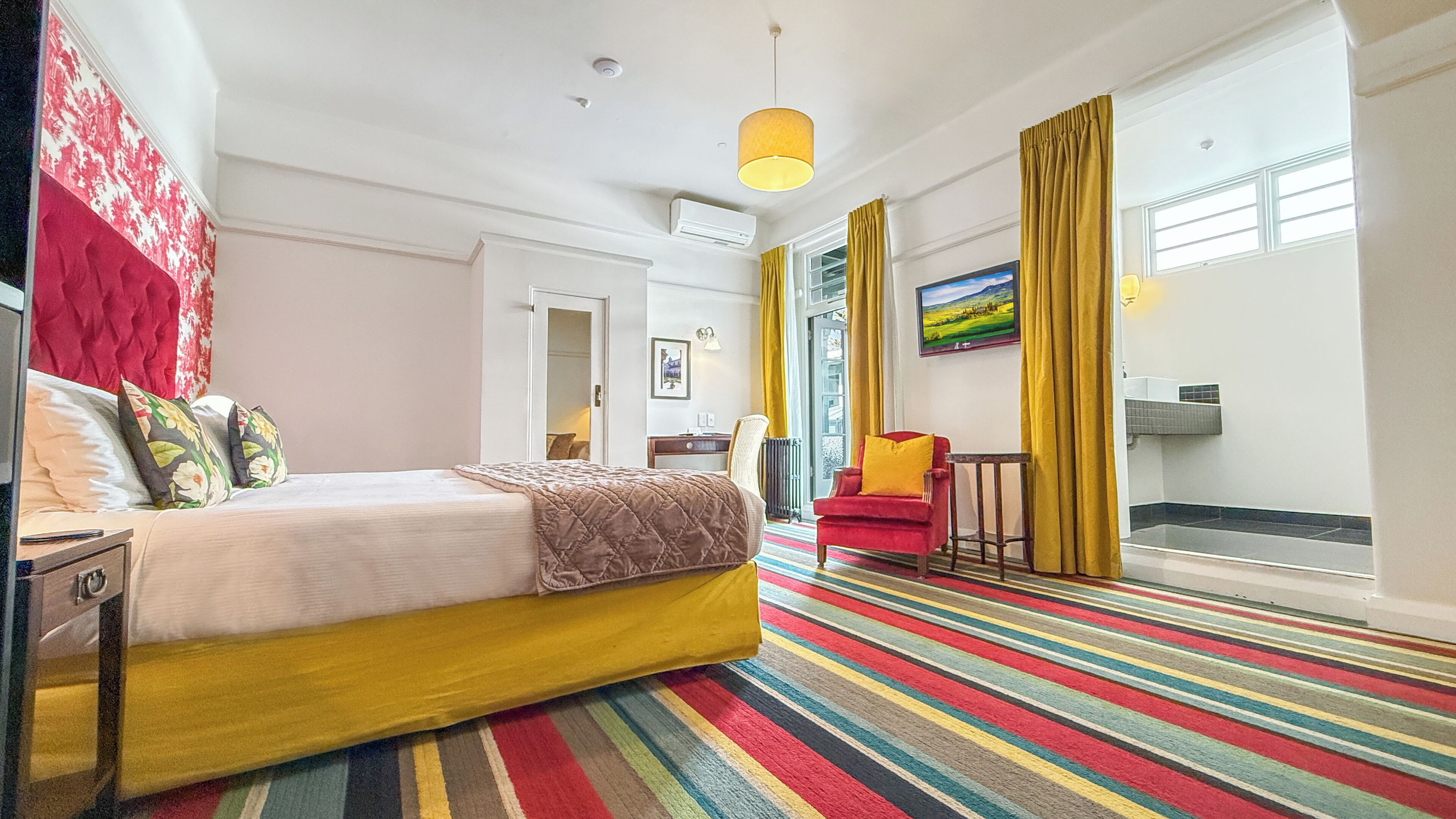 A hotel room with a bed, a desk, a chair, a colorful striped carpet, and a large window with curtains.