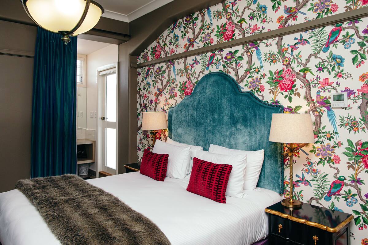 Quarto de hotel com cama, duas luminárias, cabeceira e papel de parede floral.