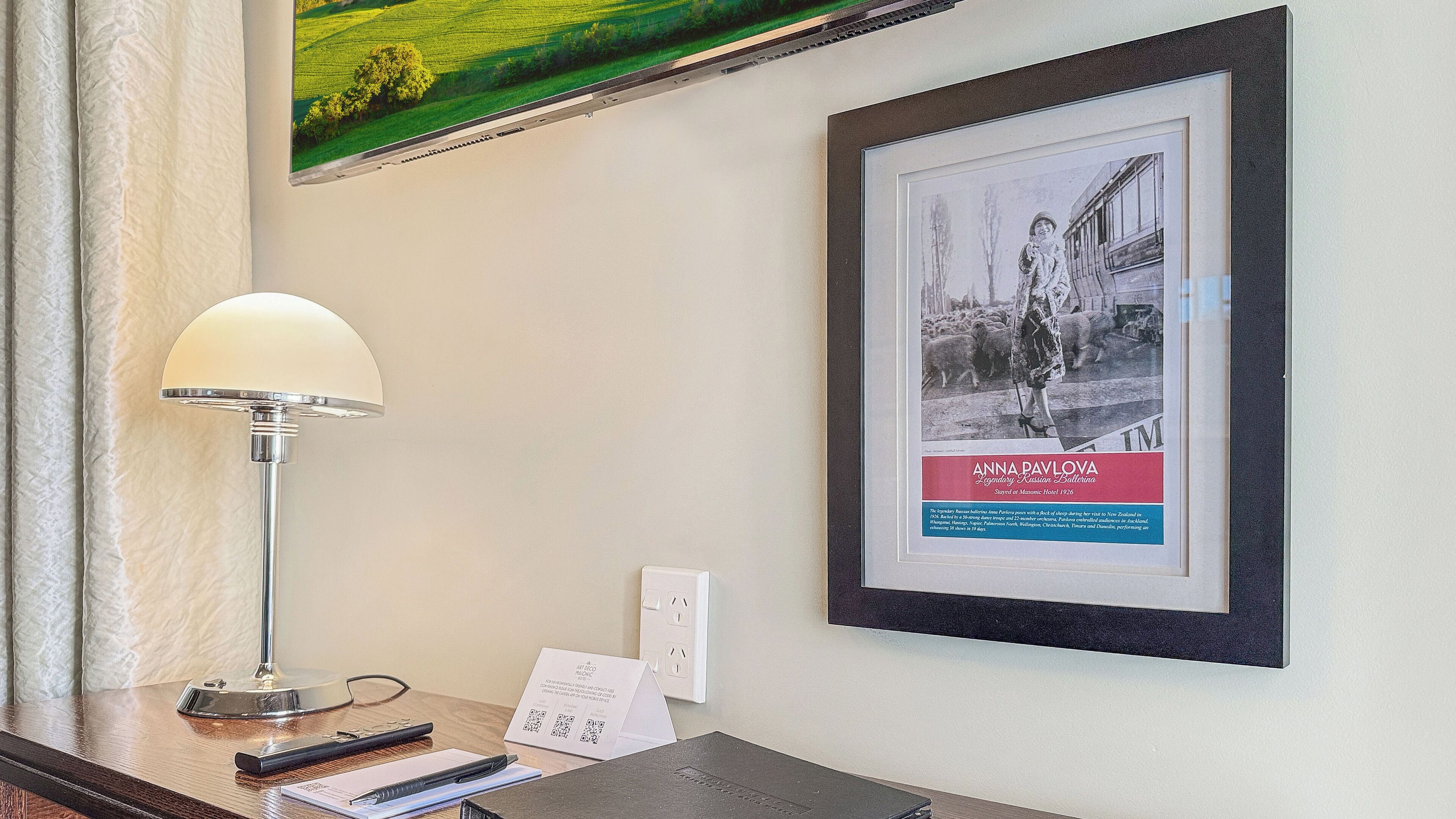 En hotellreception med ett inramat foto av Anna Pavlova, en inramad tavla ovanför disken, en lampa och ett anteckningsblock.