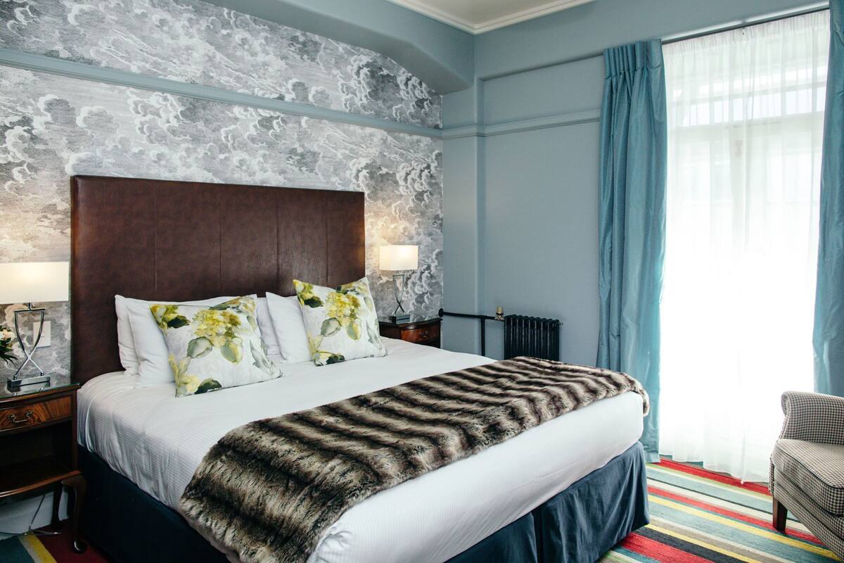 A hotel room with a large bed, floral pillows, a fur throw, a nightstand with a lamp, a radiator, and a chair by the window.