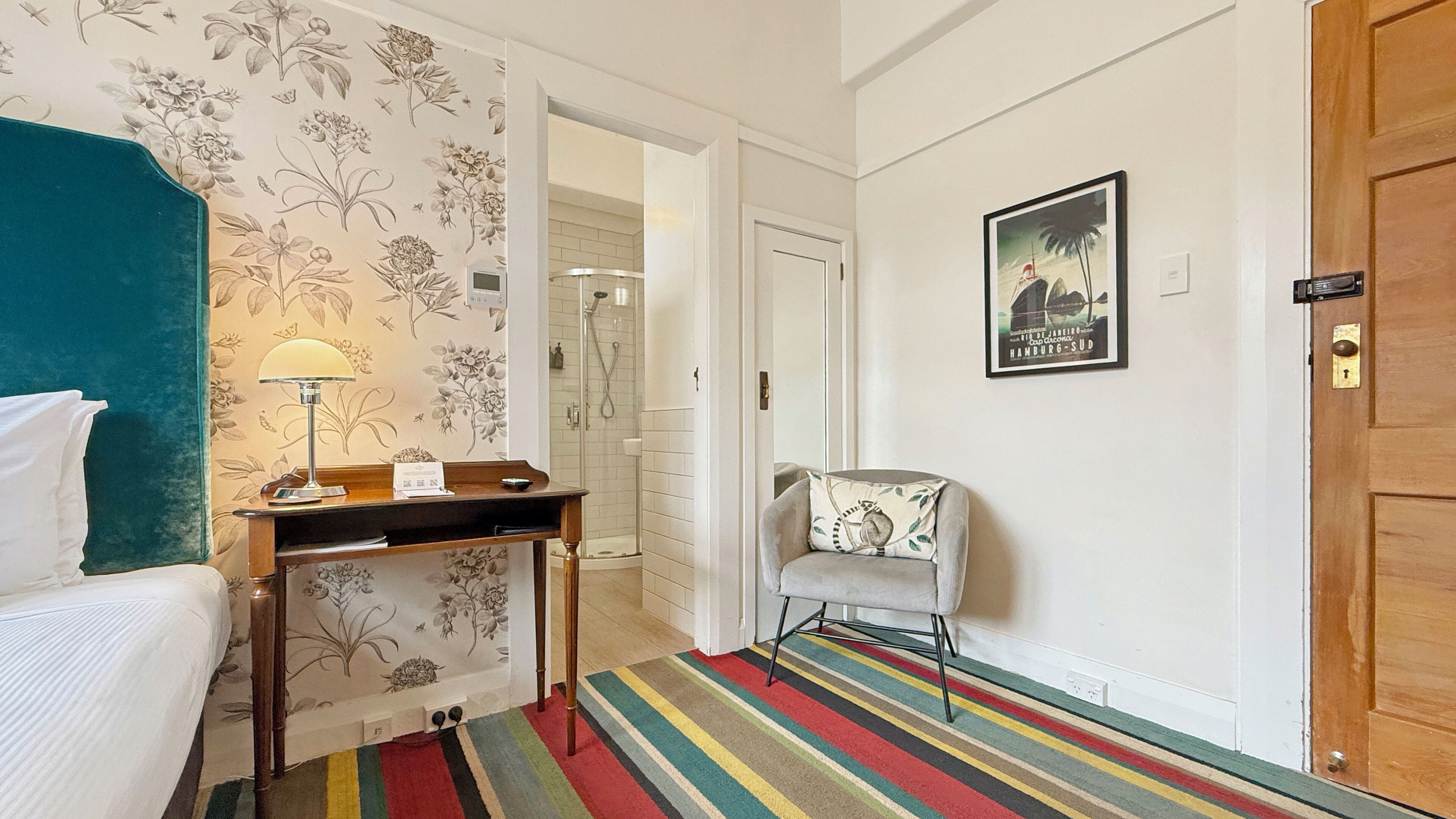 A hotel room with a bed, a desk with a lamp, a chair, a colorful striped rug, and a framed picture on the wall.