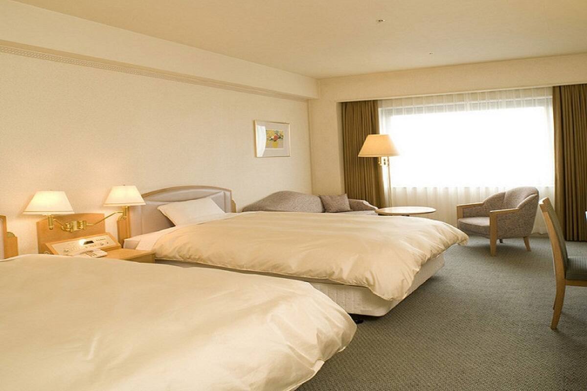 A hotel room with two beds, a chair, a desk with a lamp, and a window with curtains.