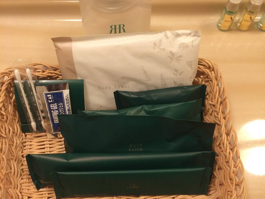 A wicker basket containing a body towel, shaving gel, razors, and green bags, likely toiletries, on a hotel counter.
