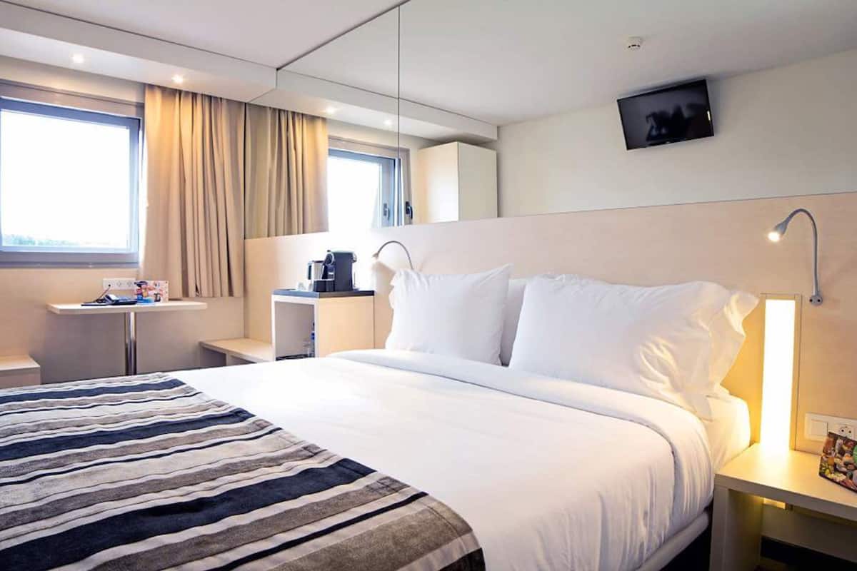 Habitación de hotel con cama, mesita de noche, lámpara, televisión y ventana con cortinas.