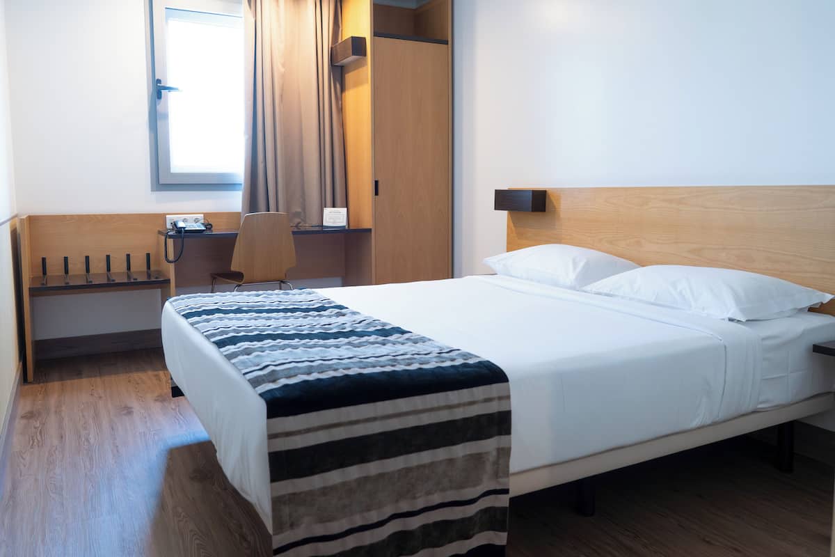 Habitación de hotel con cama, escritorio, silla, armario y ventana con cortinas.