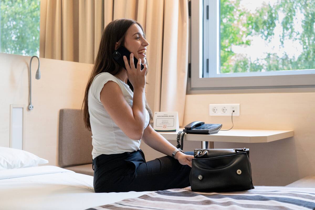 Habitación de hotel con una cama, un escritorio con una computadora, un teléfono y un bolso.