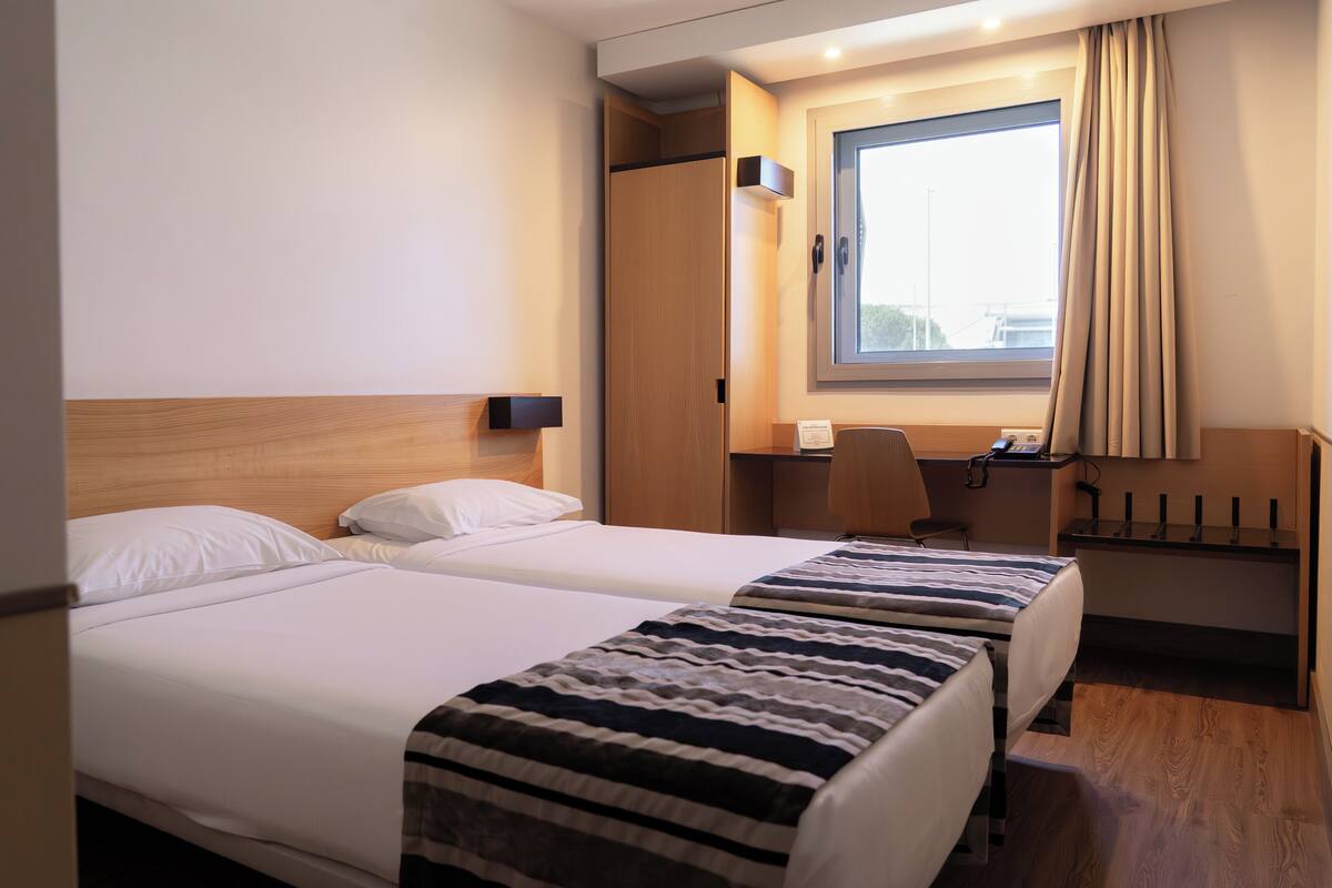 Habitación de hotel con dos camas, un escritorio, una silla, un ventanal con cortinas y suelo de madera.