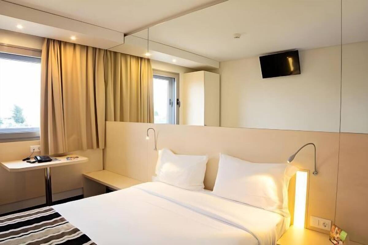 Habitación de hotel con cama, mesita de noche, lámpara, televisión y ventana con cortinas.