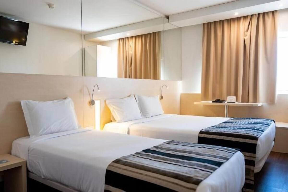 Habitación de hotel con dos camas, un televisor montado en la pared, una mesita de noche y una ventana con cortinas.