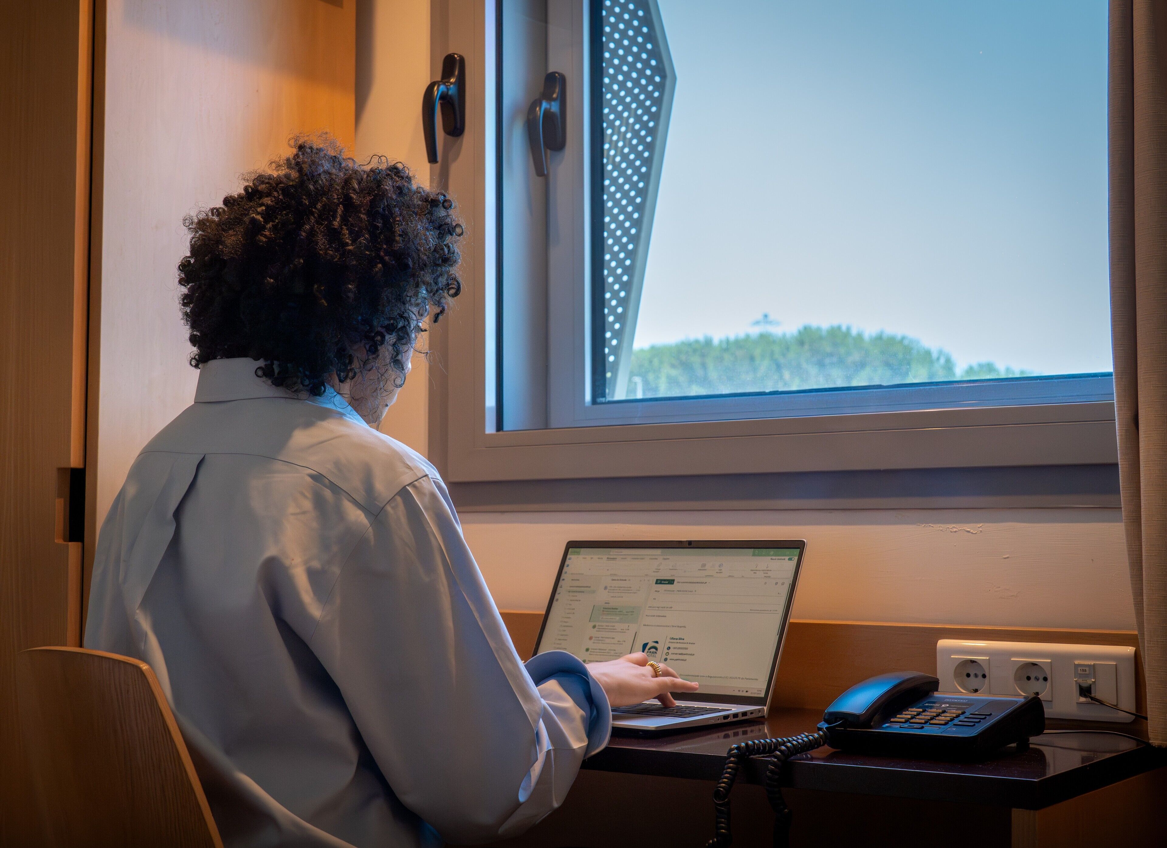 A person working at a desk with a laptop, a telephone, and a window with a view of trees.