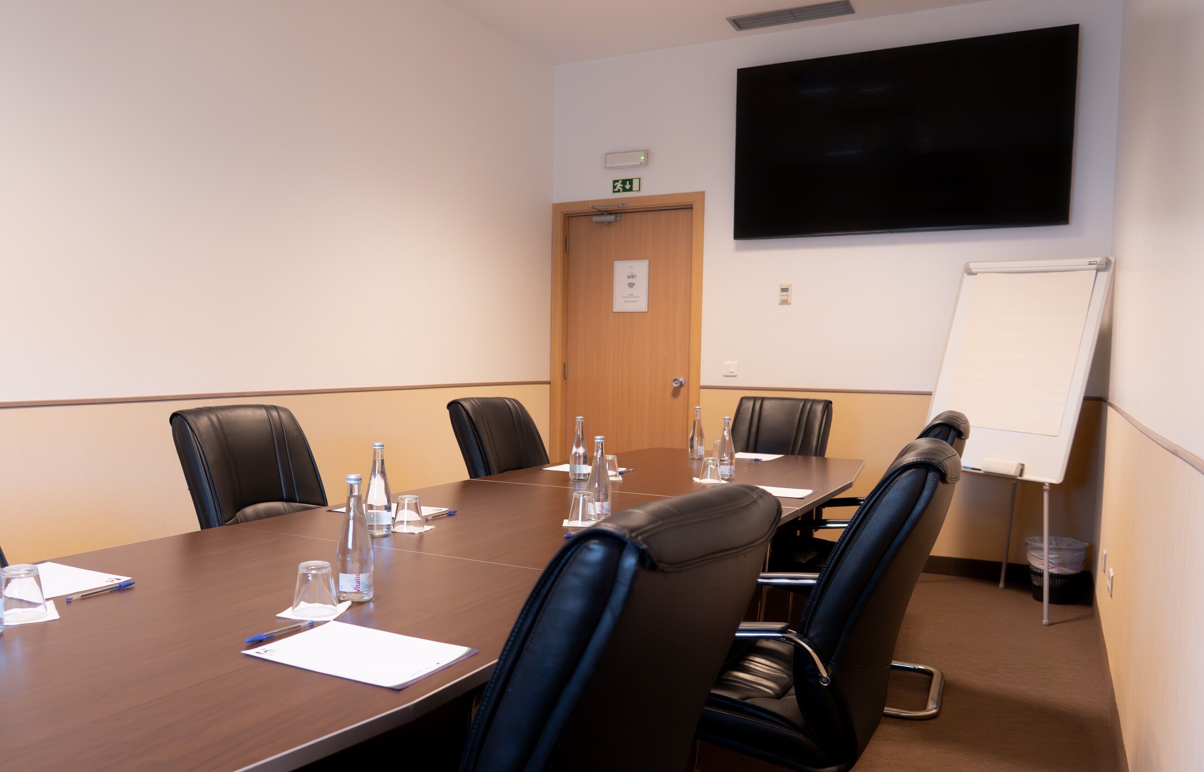 A conference room with a long table, chairs, water bottles, papers, a whiteboard, and a TV on the wall.