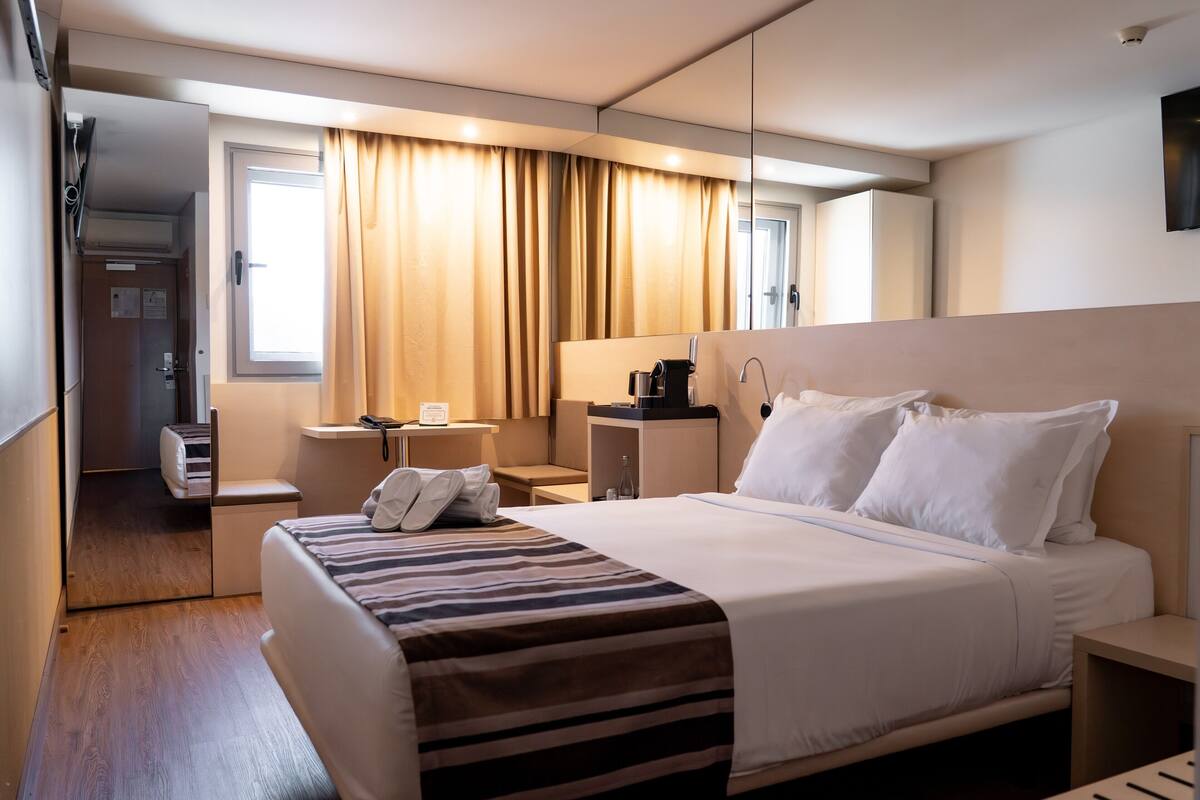 Una habitación de hotel con cama, mesitas de noche, un espejo, una ventana con cortinas y una puerta que da al baño.